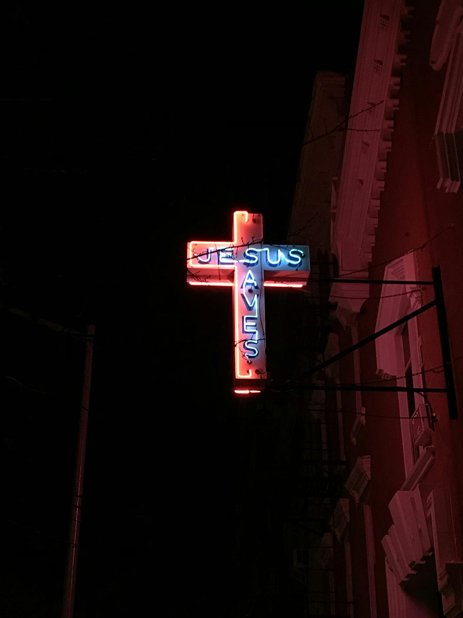 A glowing neon cross sign that reads 'Jesus Saves' against a dark night sky.